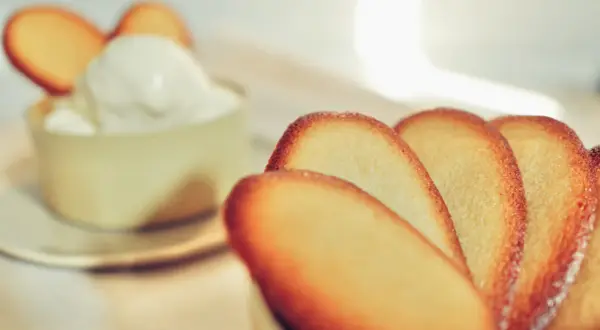 Biscotti croccanti a forma di lingue di gatto, alcuni in primo piano, altri decorano una coppa di panna montata.