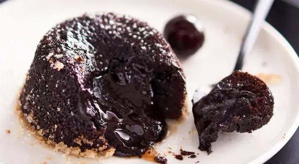 Un delizioso tortino al cioccolato con cuore fuso, cosparso di zucchero a velo, su un piatto bianco con una ciliegia e un cucchiaino.