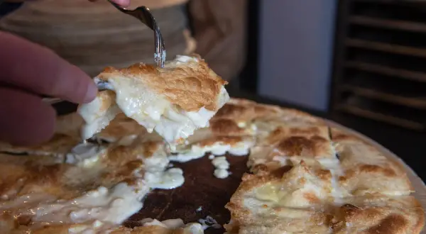 Una fetta di focaccia di Recco ricetta, filante e dorata, viene sollevata con una forchetta.