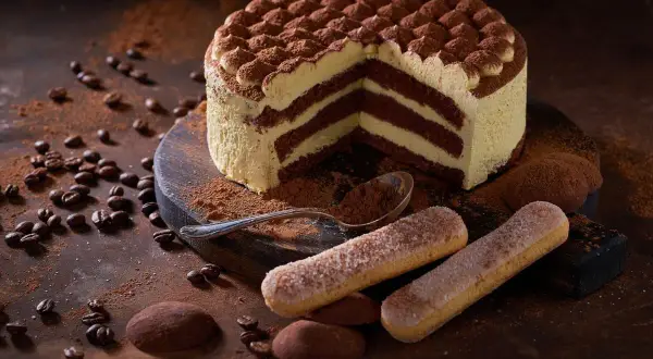 Una fetta di torta tiramisù, con strati di crema e cacao, circondata da savoiardi, chicchi di caffè e cacao in polvere.