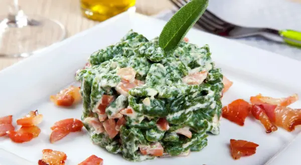 Spätzle verdi cremosi con pezzetti di speck croccante, guarniti con una foglia di salvia. Un piatto invitante.