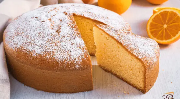 Torta soffice all'arancia, ricetta della nonna, cosparsa di zucchero a velo, con fette d'arancia sullo sfondo.