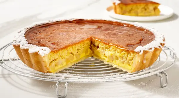 Torta di mele con crema pasticcera, con una fetta tagliata, cosparsa di zucchero a velo su una griglia metallica.
