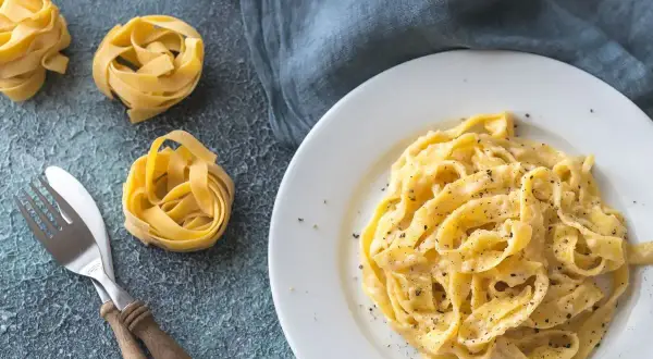 Un piatto di fettuccine alfredo cremoso, con nidi di pasta cruda e posate sul lato.