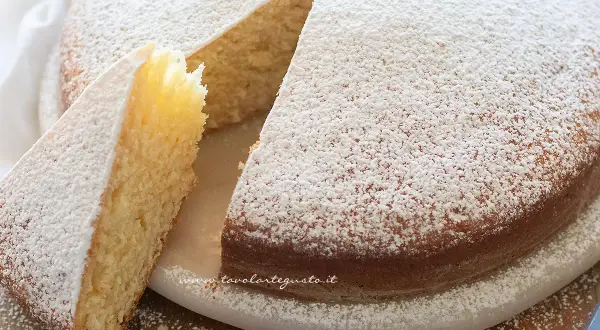 Una soffice torta senza uova, cosparsa di zucchero a velo, con una fetta tagliata che ne mostra l'interno chiaro e profumato.