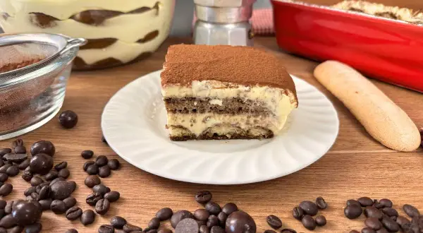Una fetta di tiramisù con uova pastorizzate, cosparso di cacao, su un piatto bianco. Intorno, chicchi di caffè, palline di cioccolato e un savoiardo.