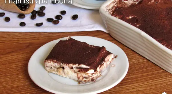 Una fetta di tiramisù alla ricotta, cosparso di cacao, su un piatto bianco, con chicchi di caffè e una teglia sullo sfondo.