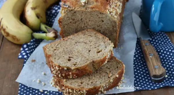 Fetta di banana bread senza burro, soffice e profumato, con crosta croccante di frutta secca. Accanto, banane mature e una tazza blu.