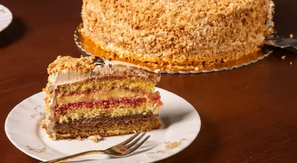 Una fetta di pizza dolce abruzzese, multistrato e ricoperta di nocciole, su un piatto bianco con una forchetta dorata.