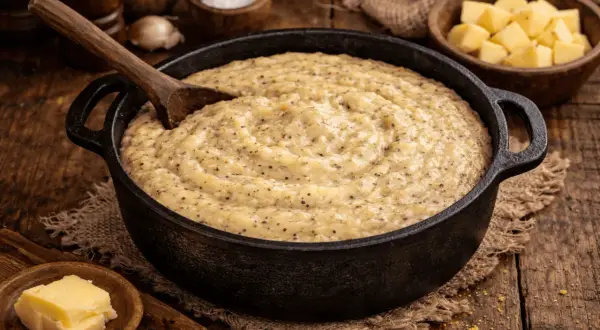 Una calda polenta taragna fumante in una padella di ghisa, con un cucchiaio di legno pronto a servirla. Accanto, cubetti di formaggio e burro.