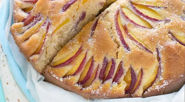 Una fetta di torta di pesche morbidissima, con pesche affettate disposte a raggiera sulla superficie dorata e cosparsa di zucchero a velo.