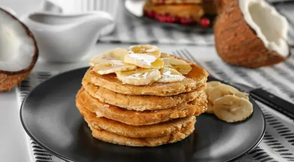Pancakes con farina di cocco, guarniti con banane a fette e sciroppo, su un piatto nero. A lato, un cocco aperto.