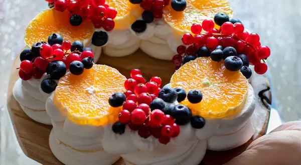 Torta pavlova a forma di corona, decorata con fette d'arancia, ribes rossi e mirtilli.