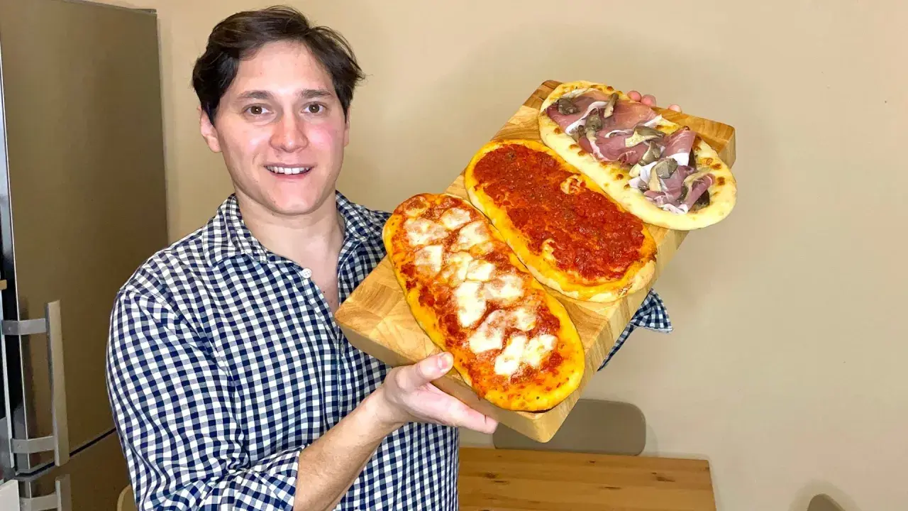 Uomo sorridente mostra tre lingue di pizza senza lievito: una margherita, una al pomodoro e una con prosciutto e funghi.