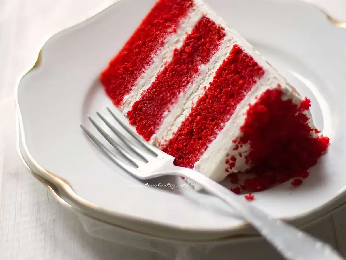 Una fetta di torta red velvet, stratificata con crema bianca, su un piatto bianco con una forchetta.