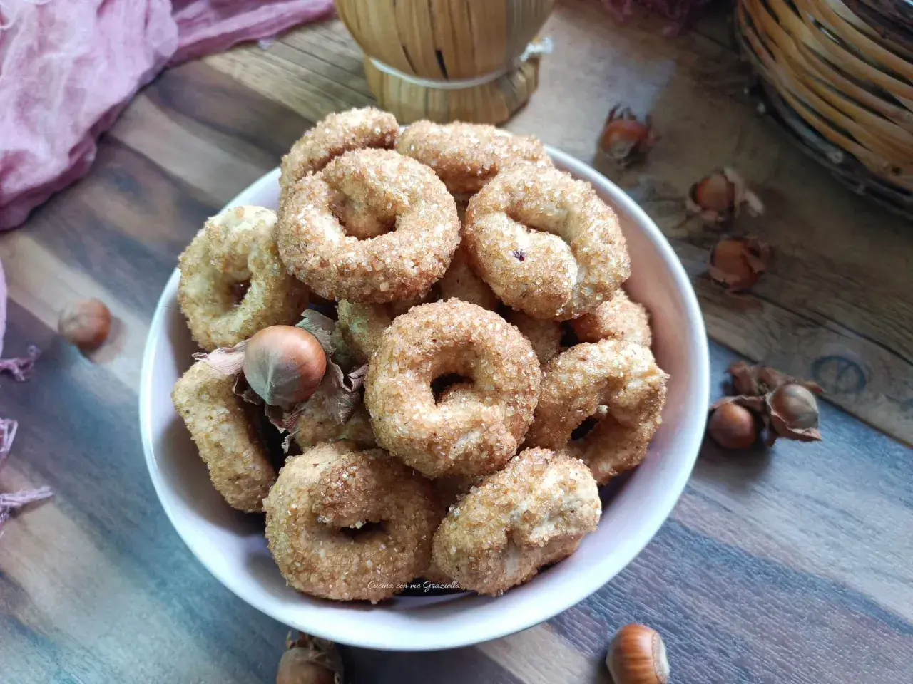 Ciambelline al vino cosparse di zucchero, con nocciole e un drappo rosa sullo sfondo.