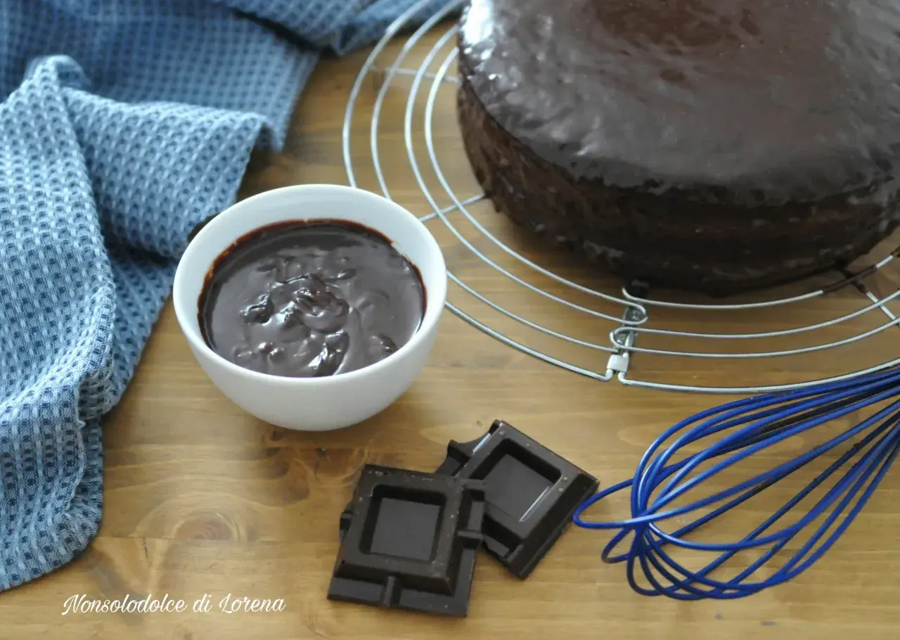 Torta ricoperta di lucida ganache cioccolato per copertura, con ciotolina di ganache, cioccolato fondente e frusta blu.