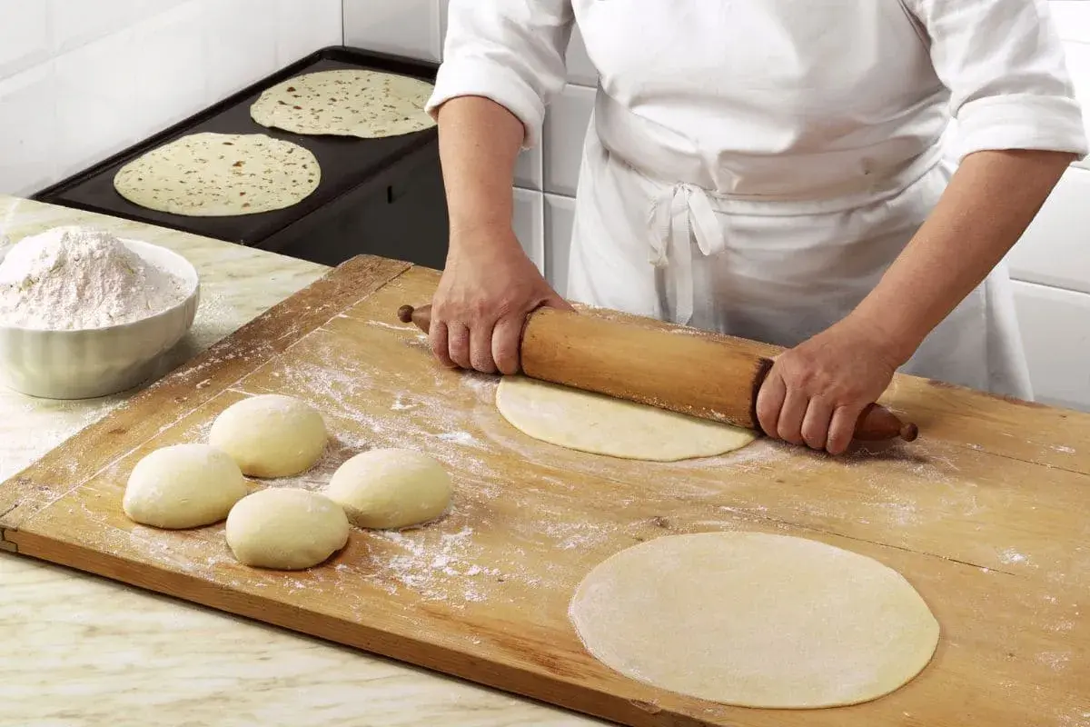 Mani esperte stendono la pasta per una piadina fatta in casa, mentre altre sono già pronte per essere cotte.