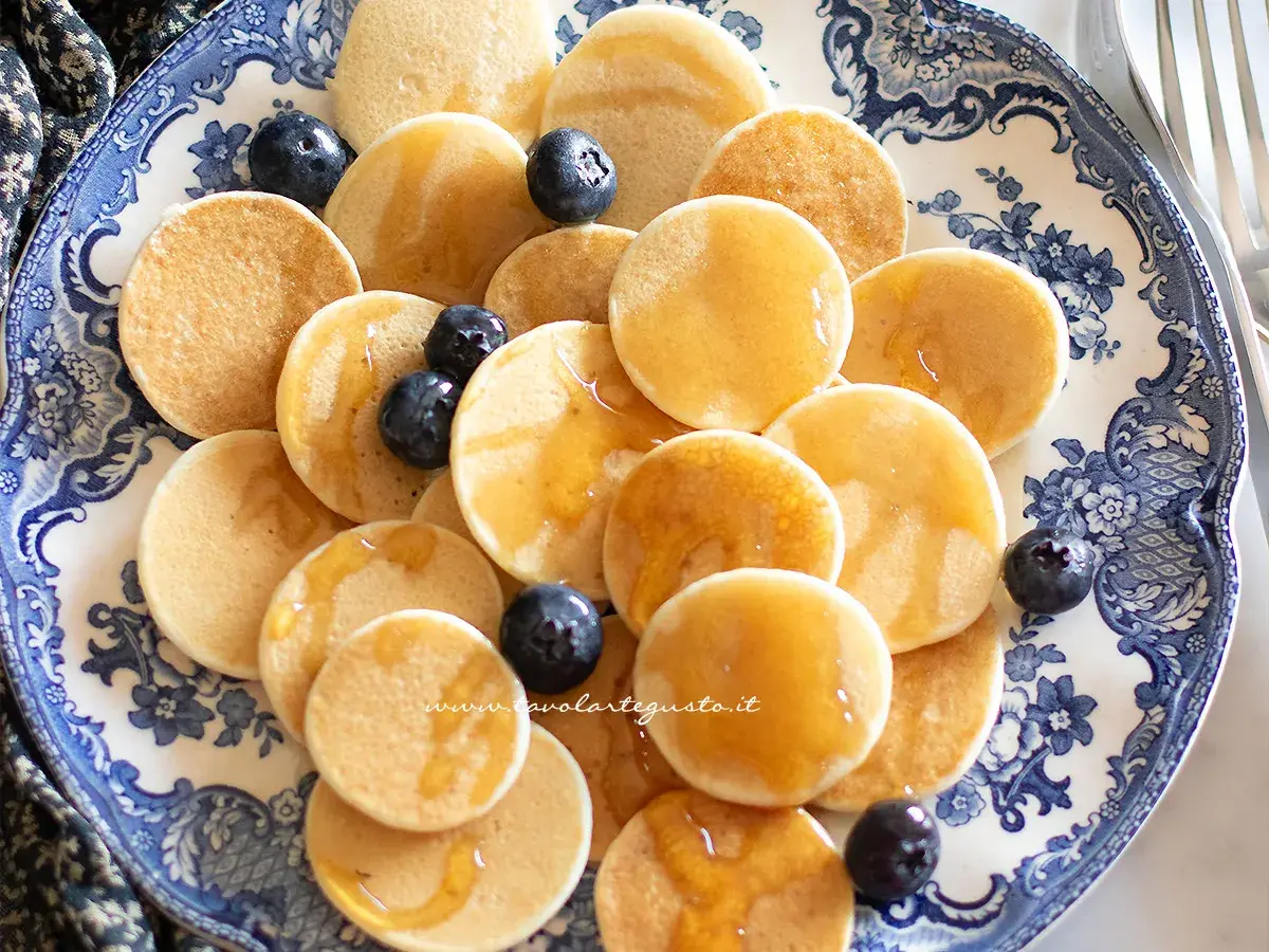 Mini pancake ricetta: tanti piccoli pancake dorati, guarniti con mirtilli freschi e sciroppo d'acero, serviti su un piatto decorato.
