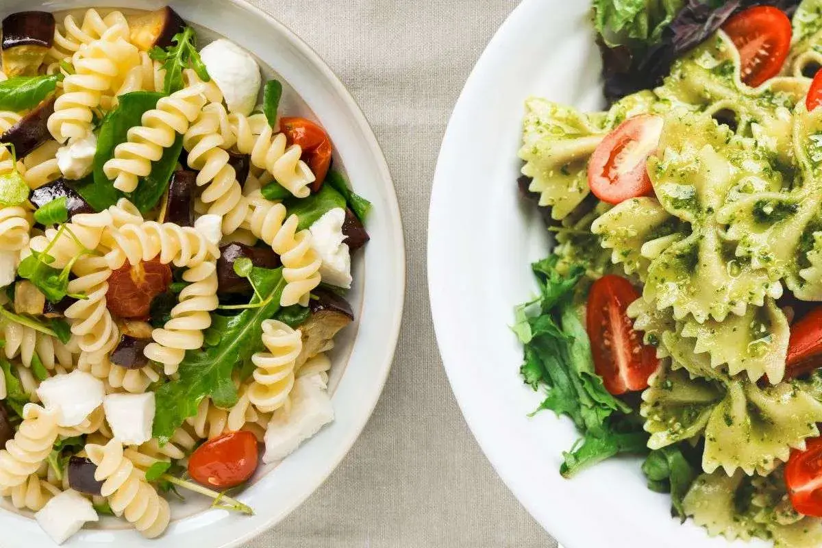 Due insalate di pasta fredda senza pomodori: una con fusilli, melanzane e mozzarella, l'altra con farfalle al pesto e pomodorini.