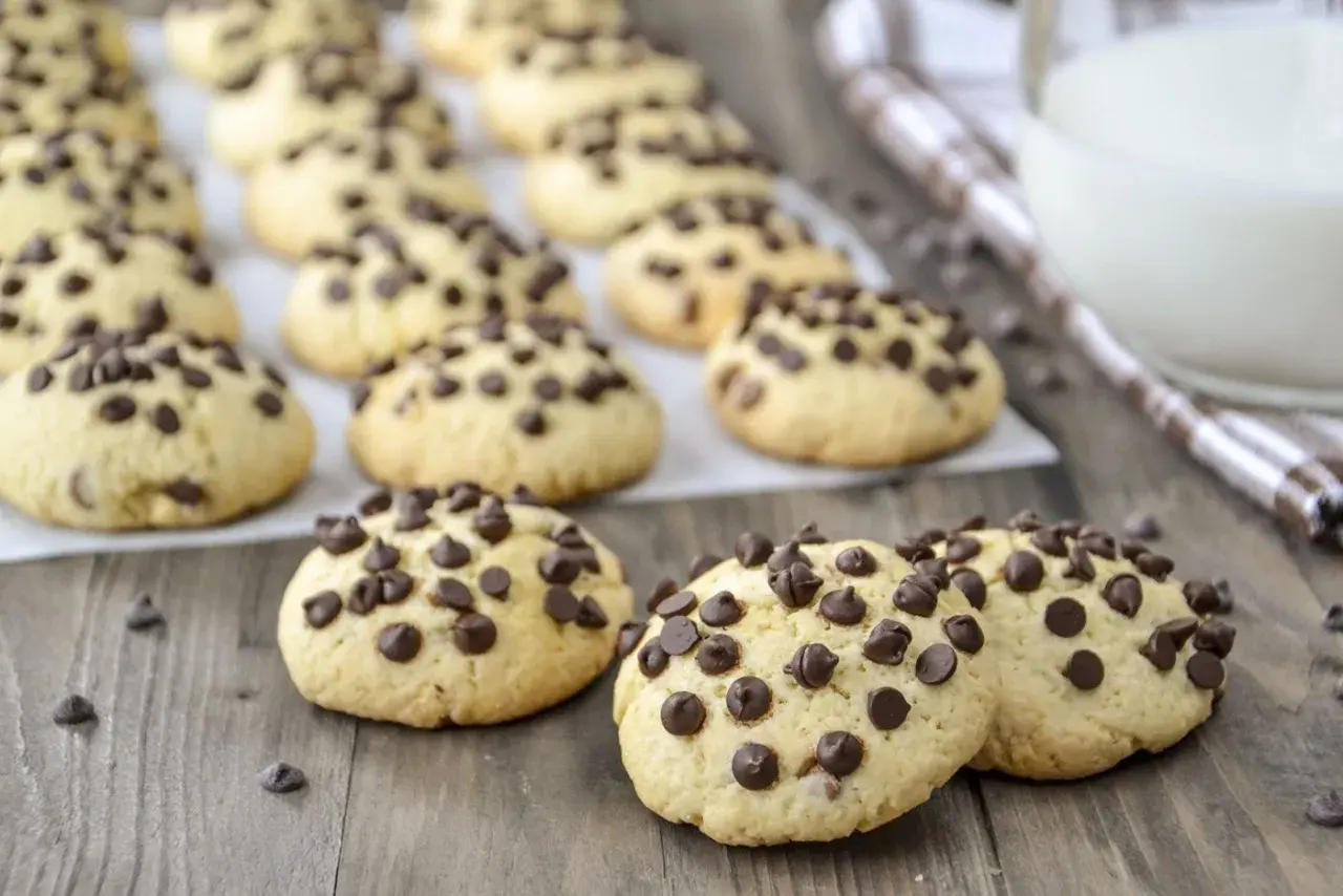 Biscotti con gocce di cioccolato, dorati e invitanti, sparsi su un tavolo di legno. Alcuni sono in primo piano, altri sullo sfondo.