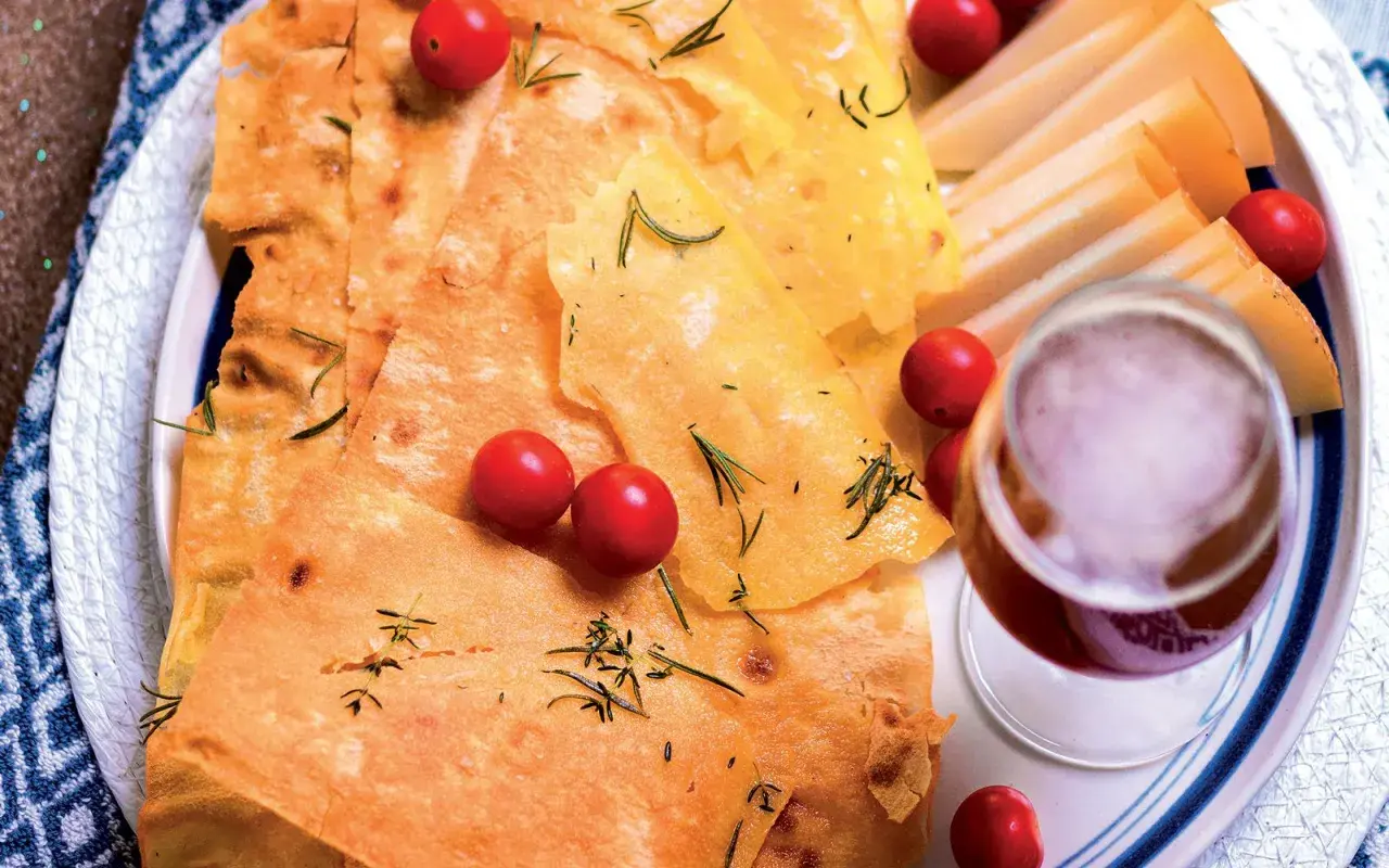Pane guttiau croccante con pomodorini, rosmarino e fette di formaggio, accompagnato da un bicchiere di vino rosso.