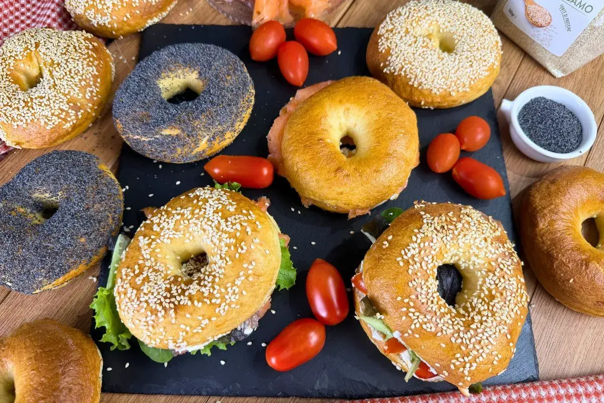 Varietà di bagel, alcuni con semi di sesamo, altri con semi di papavero, pronti per una deliziosa bagel ricetta. Pomodorini e salmone completano la scena.