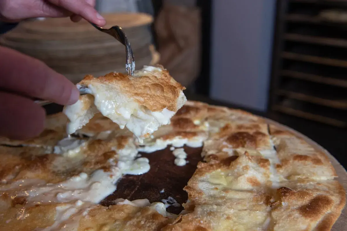 Una fetta di focaccia di Recco ricetta, filante e dorata, viene sollevata con una forchetta.