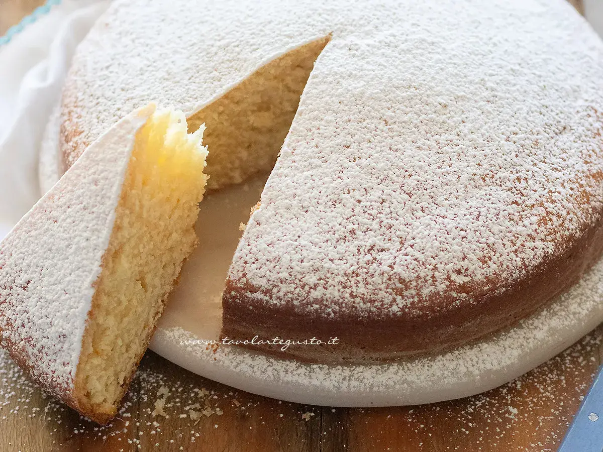 Una soffice torta senza uova, cosparsa di zucchero a velo, con una fetta tagliata che ne mostra l'interno chiaro e profumato.