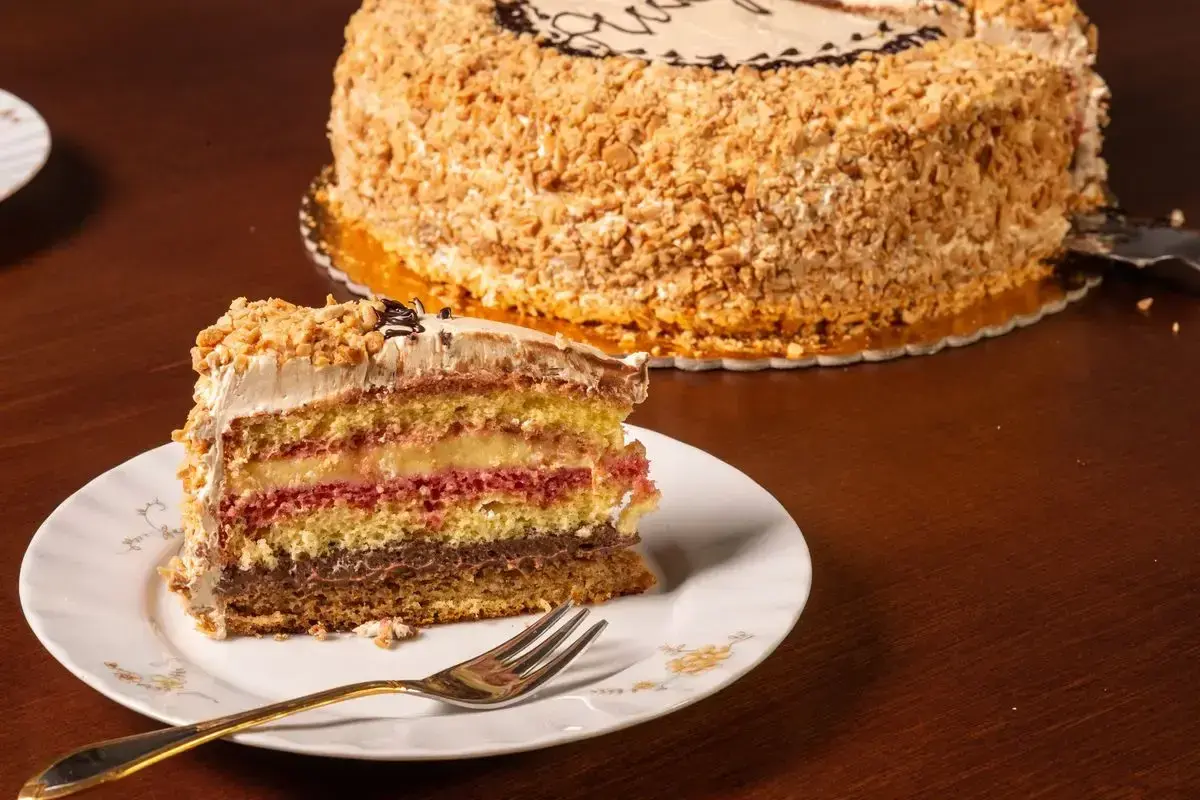 Una fetta di pizza dolce abruzzese, multistrato e ricoperta di nocciole, su un piatto bianco con una forchetta dorata.