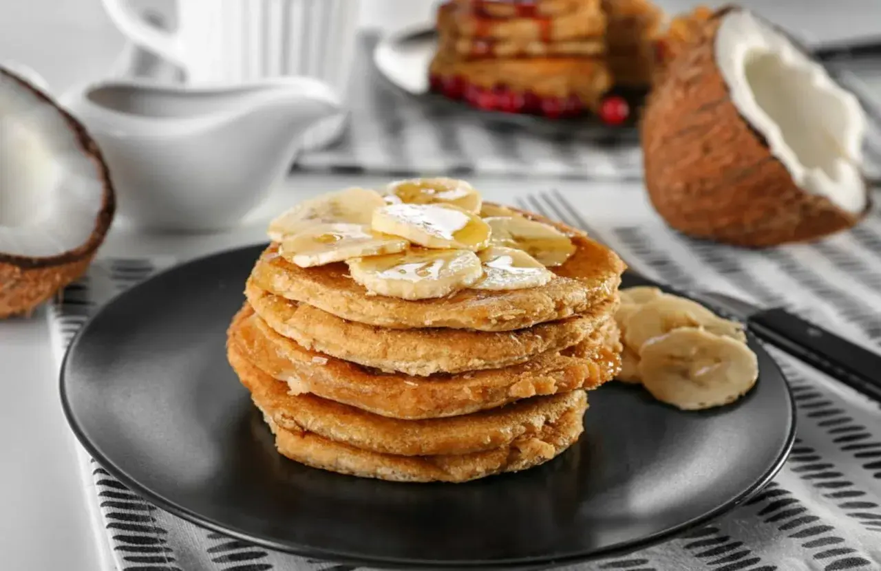 Pancakes con farina di cocco, guarniti con banane a fette e sciroppo, su un piatto nero. A lato, un cocco aperto.