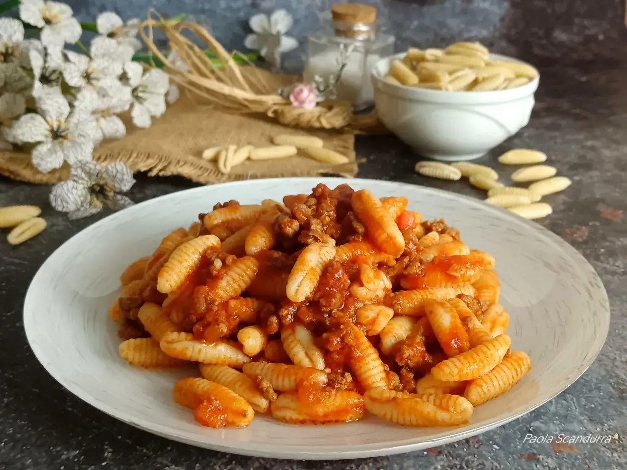 Piatto di malloreddus alla campidanese, pasta tipica sarda, conditi con sugo di carne e pomodoro, su un tavolo scuro con fiori e sale.