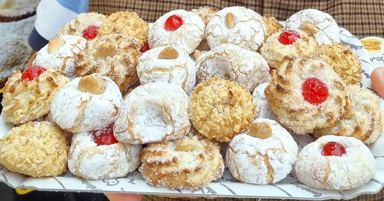 Un vassoio pieno di dolcetti fatti in casa, alcuni ricoperti di zucchero a velo e mandorle tritate, altri con ciliegie candite. Sono deliziose paste di mandorla.