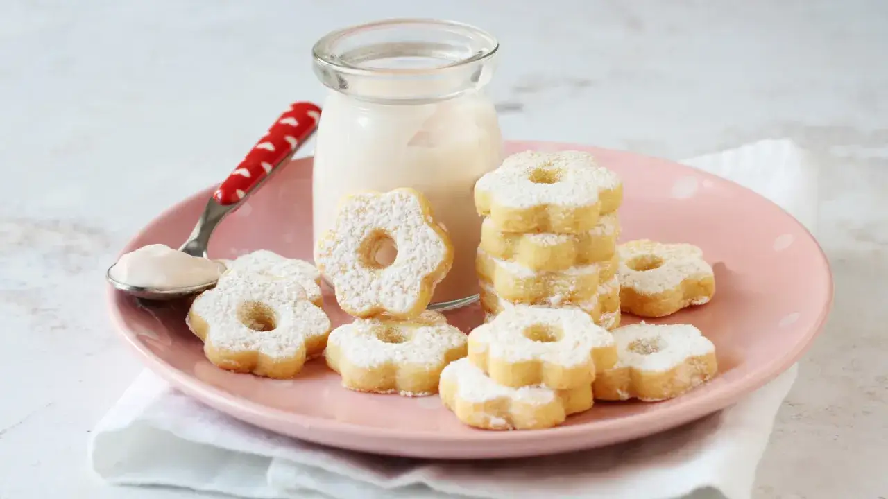 Deliziosi canestrelli ricetta: biscotti a forma di fiore spolverati di zucchero a velo, serviti con una crema e una bottiglietta di latte.