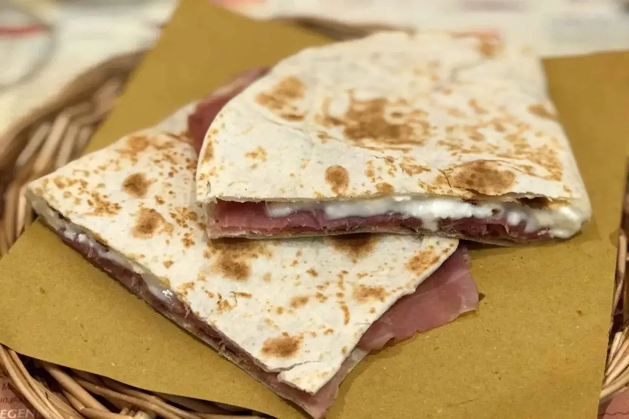 Piadina fatta in casa, ripiena di prosciutto e formaggio cremoso, servita su carta gialla in un cestino di vimini.