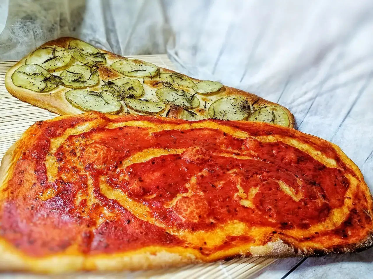 Due lingue di pizza senza lievito: una con pomodoro e l'altra con patate e rosmarino, pronte per essere gustate.