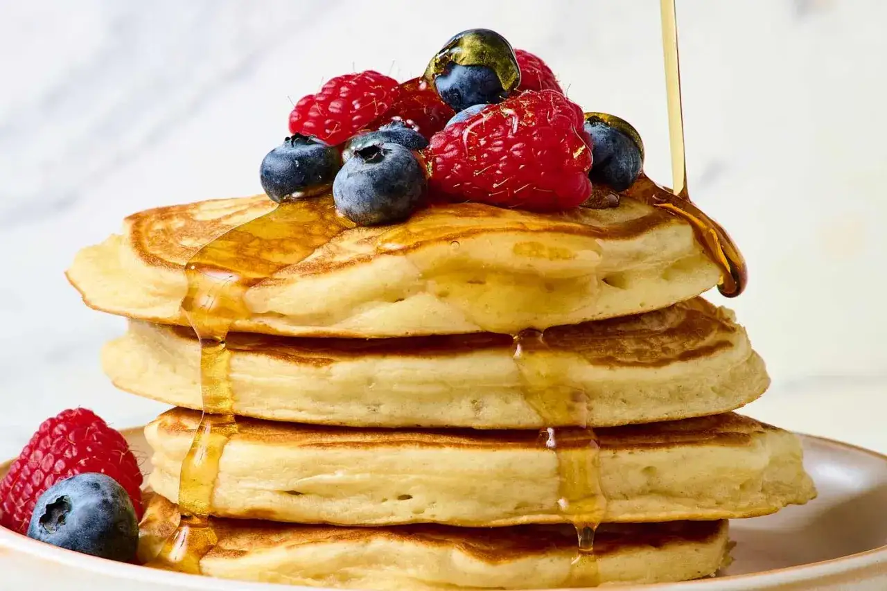 Pancakes proteici dorati, impilati e ricoperti di sciroppo e frutti di bosco freschi.