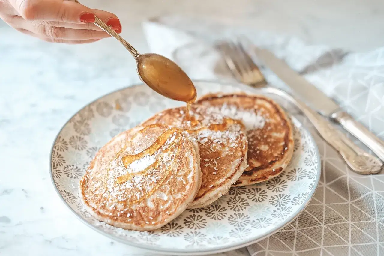 Pancake con farina di cocco, spolverati di zucchero a velo e irrorati con sciroppo dorato, pronti per una colazione golosa.