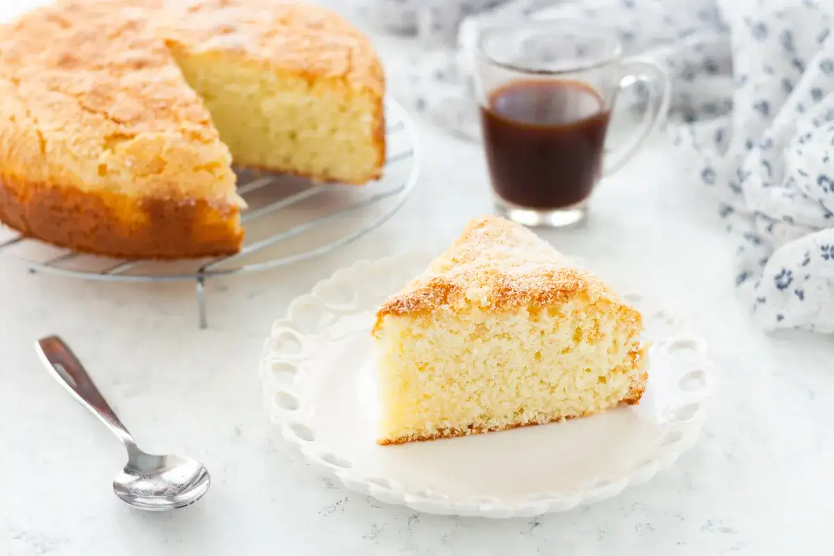 Una fetta di torta ricotta e cocco, soffice e dorata, su un piatto bianco. Accanto, una tazzina di caffè e la torta intera su una griglia.