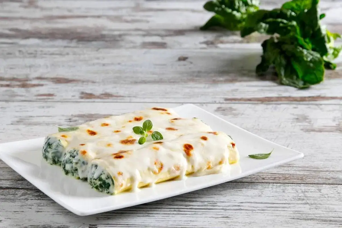 Cannelloni ricotta e spinaci gratinati su un piatto bianco, con foglie di spinaci freschi sullo sfondo.