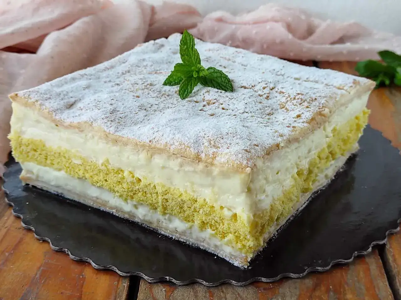 Una fetta di torta diplomatica, soffice e stratificata, con crema pasticcera e pan di Spagna, spolverata di zucchero a velo e guarnita con menta.