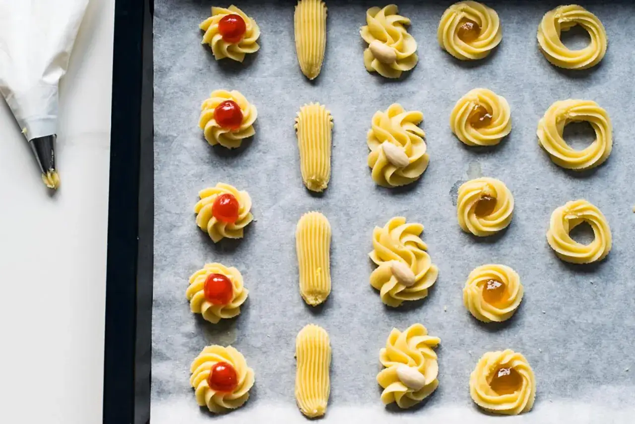 Biscotti viennesi ricetta originale: dolcetti su teglia pronti per la cottura, alcuni decorati con ciliegie candite, mandorle o marmellata.