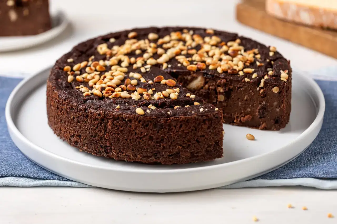 Torta paesana al cioccolato, ricca e umida, guarnita con pinoli tostati. Una fetta &egrave; stata gi&agrave; servita, invitando a gustare questo dolce rustico.