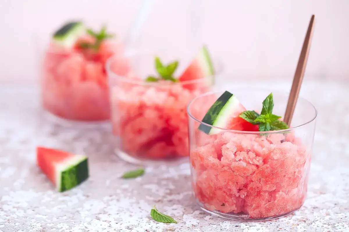 Tre bicchieri di rinfrescante granita di anguria, guarniti con spicchi di frutto e foglie di menta. Perfetta per l'estate.