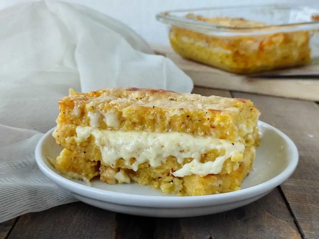 Una fetta di pasticcio di polenta dorata e filante, con formaggio fuso tra gli strati, servita su un piatto bianco.