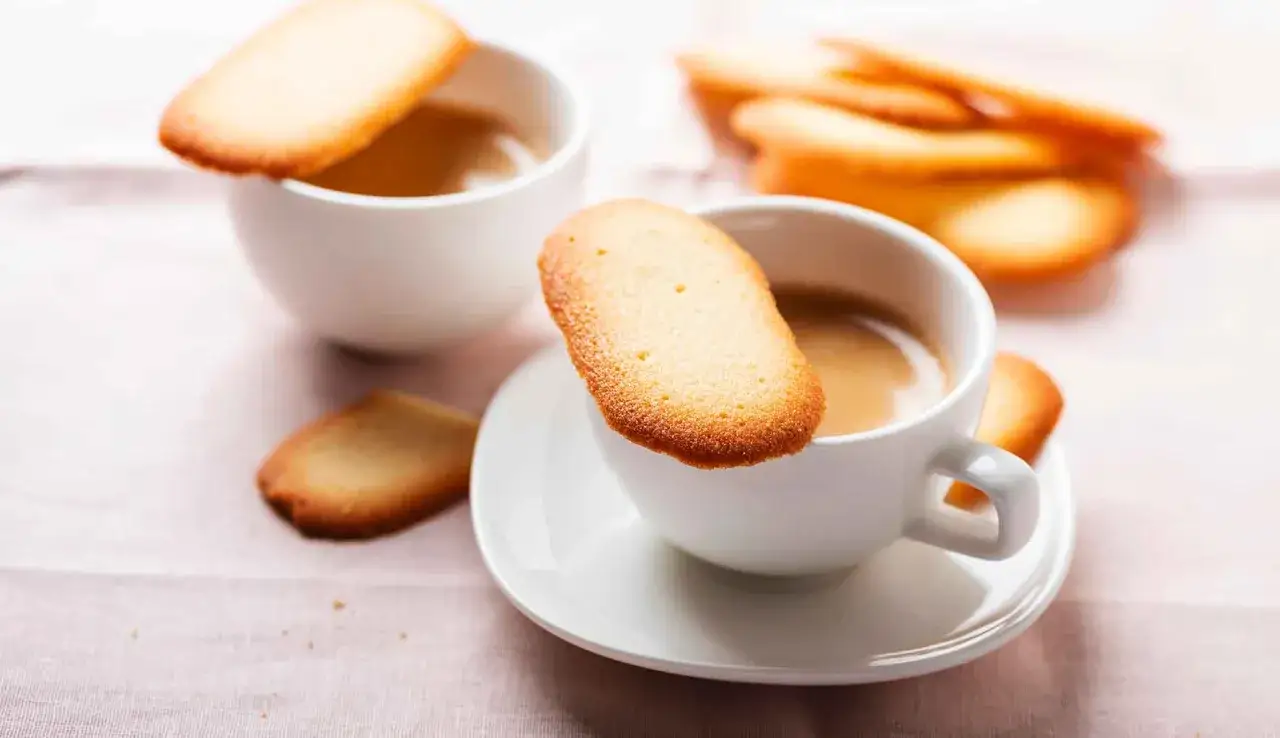 Biscotti lingue di gatto dorati appoggiati su tazze di caffè fumante, un invito alla dolcezza.