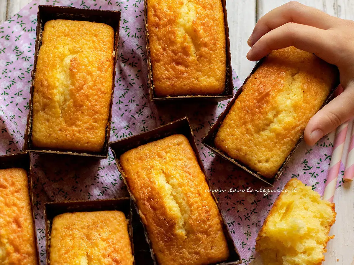 Plumcake yogurt dorati e soffici, pronti per essere gustati. Una mano ne afferra uno, mentre un pezzetto è già stato assaggiato.