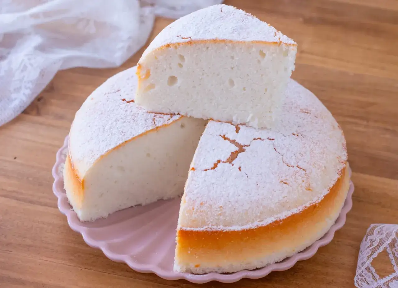 Una soffice torta nuvola, cosparsa di zucchero a velo, su un piatto rosa. Una fetta è appoggiata sopra, invitando a gustarla.