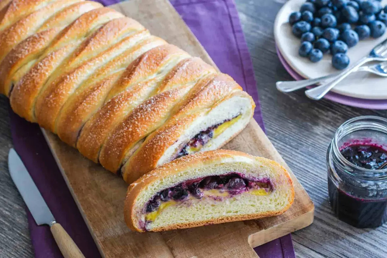 Deliziosa treccia mochena ripiena di mirtilli e crema, cosparsa di zucchero, su tagliere di legno.