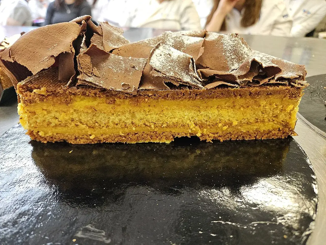 Fetta di torta pazientina con strato giallo intenso e copertura di riccioli di cioccolato fondente.